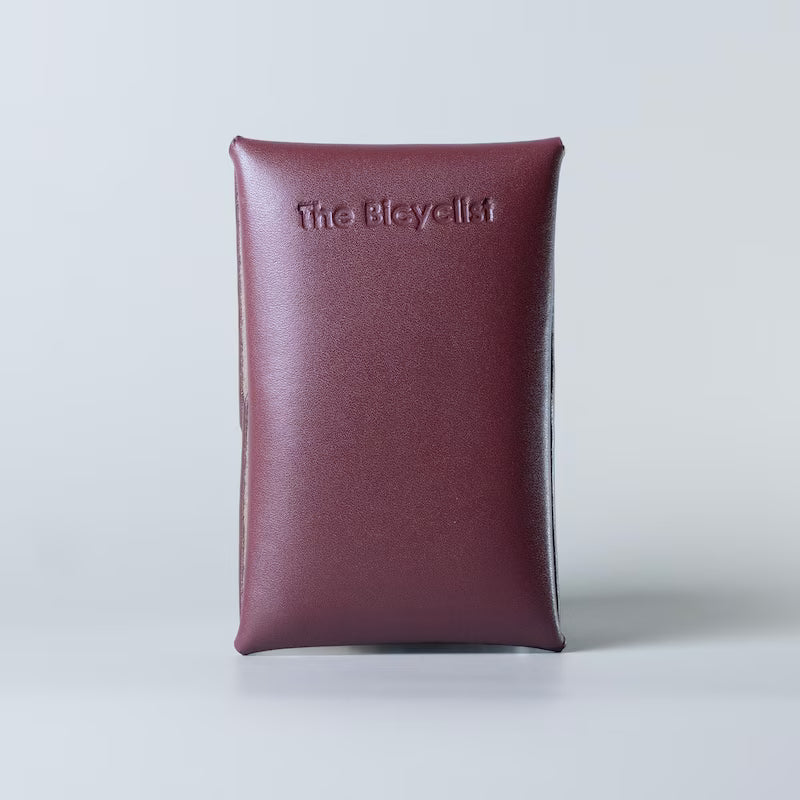 A burgundy or maroon full grain leather card holder with 'The Bicyclist' embossed on the front, set against a light gray background.