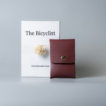 A burgundy or maroon full grain leather card holder or small card pouch with a snap button closure, displayed in front of a white box labeled 'The Bicyclist' on a light gray background.