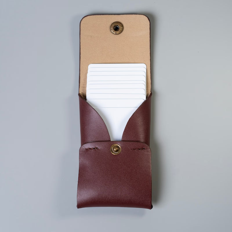 A maroon leather card holder or card wallet with a brass snap button, shown open to reveal a stack of white plastic cards inside, made by 'The Bicyclist' set against a plain light gray background.