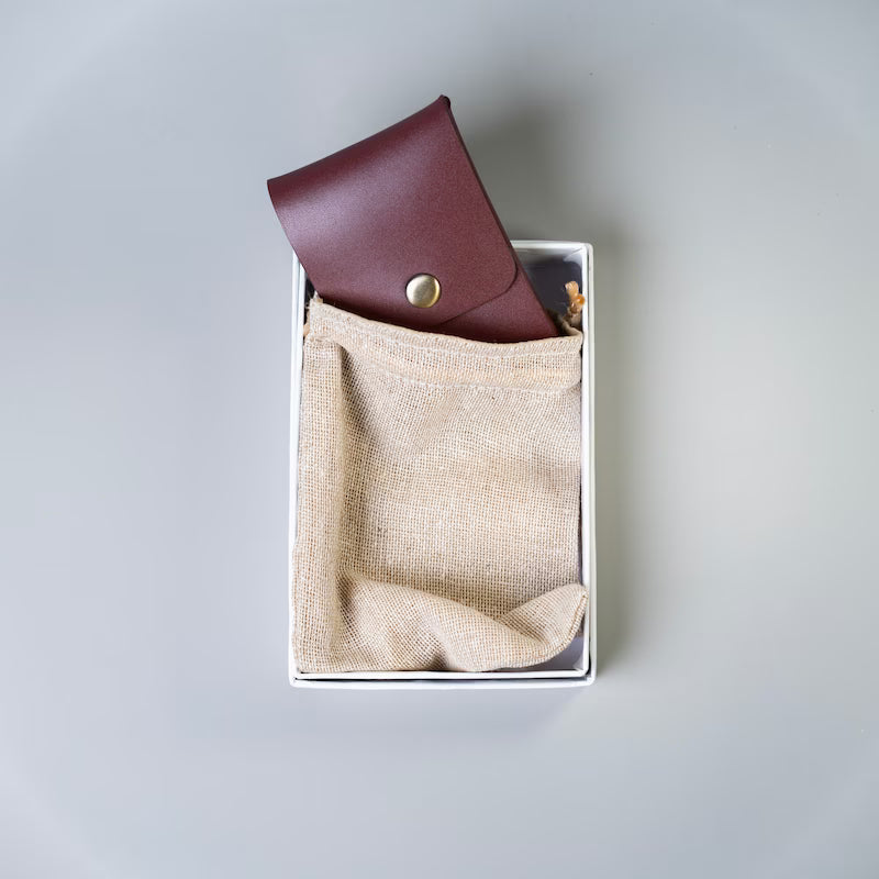 A burgundy or maroon leather card holder with a gold snap button, made by 'The Bicyclist' partially tucked into a beige jute dust bag inside a white gift box, set against a neutral gray background.