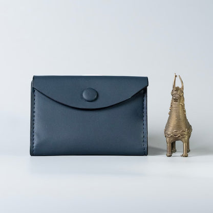 A dark navy blue full grain leather envelope-style card holder wallet with a snap button closure, made by 'The Bicyclist' shown next to a small brass horse figurine on a light gray background.