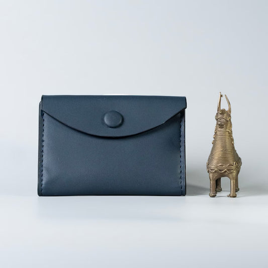 A dark navy blue full grain leather envelope-style card holder wallet with a snap button closure, made by 'The Bicyclist' shown next to a small brass horse figurine on a light gray background.