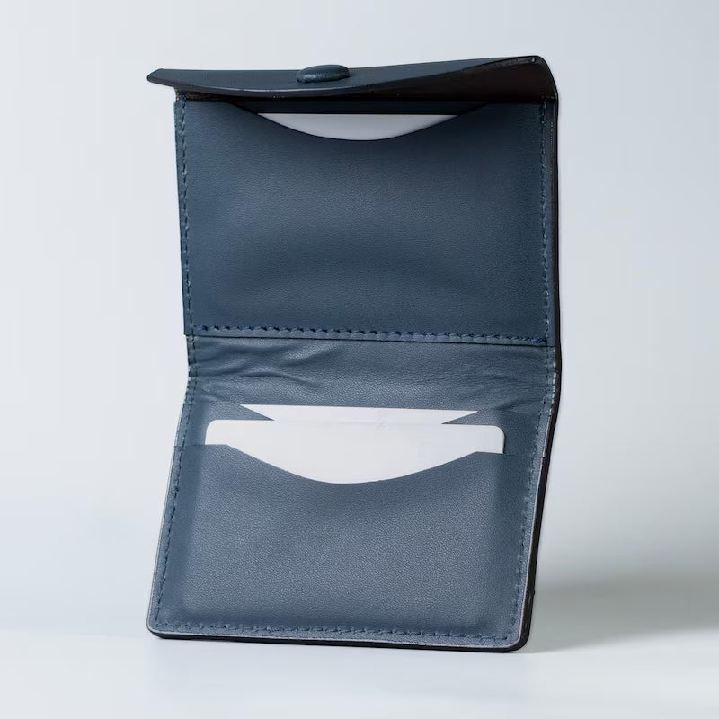 A dark blue full grain leather card holder wallet shown open, displaying its interior stitching, button closure, and card slots with white plastic cards inserted into the card slots against a light gray background.