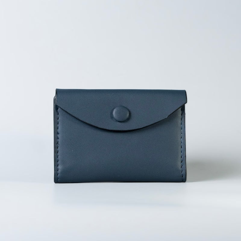 A minimalist navy blue full grain leather card holder with an envelope-style flap and a single round snap button, made by The Bicyclist photographed on a clean light gray background.