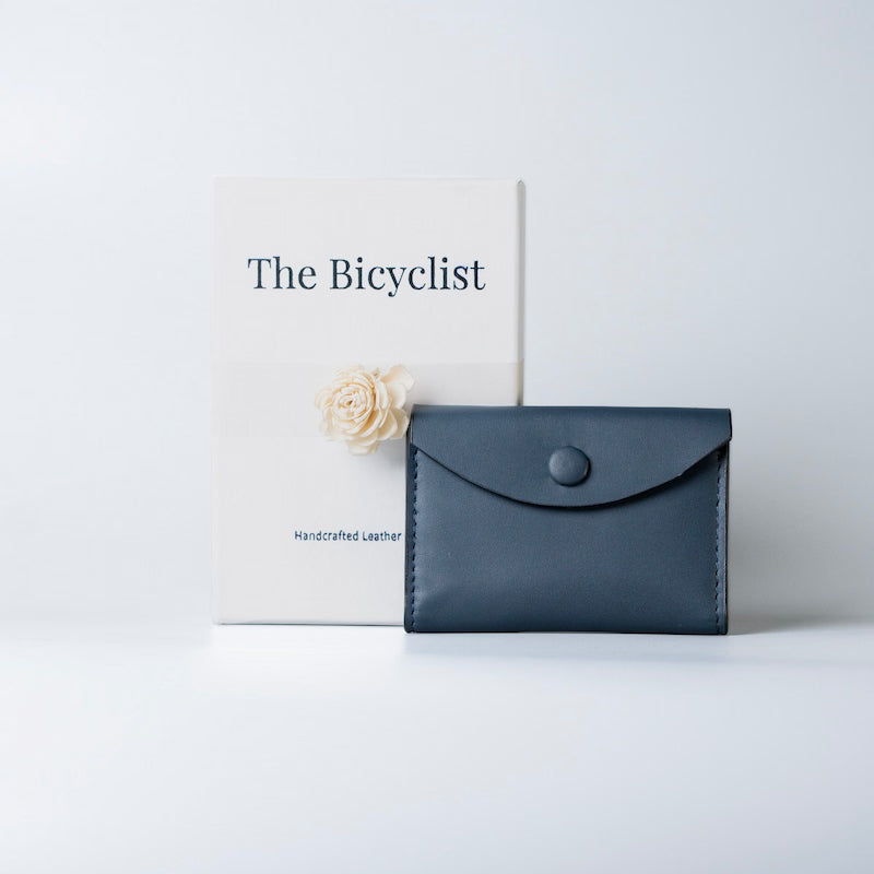 A navy blue leather envelope-style wallet with a snap button closure, displayed next to a white box with the branding 'The Bicyclist' on a clean white background.