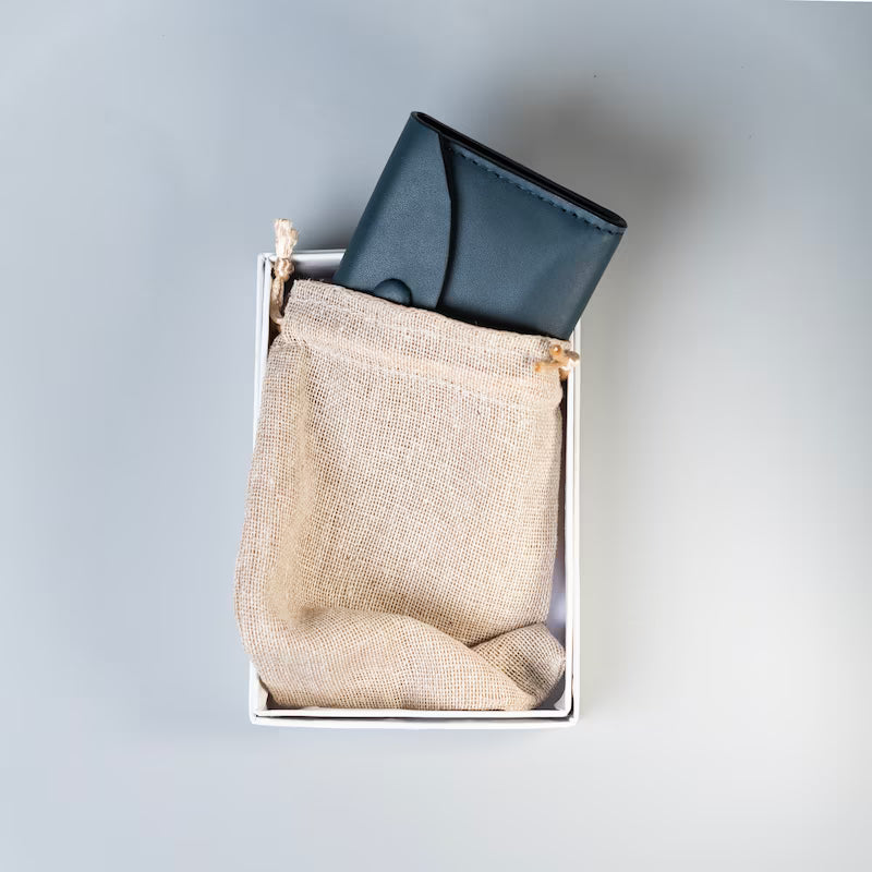 A navy blue leather wallet tucked inside a white gift box with a beige burlap drawstring pouch underneath, shot from a top-down angle on a light gray background.