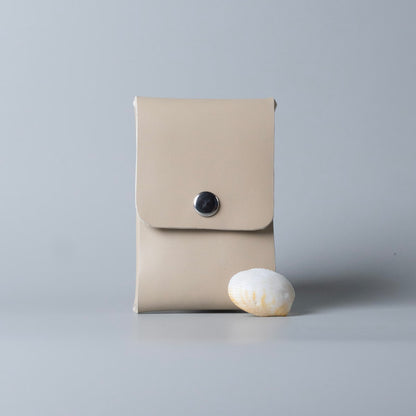 A minimalist off white full grain leather card pouch with a flap closure and a single black snap button from The Bicyclist, standing upright on a light gray surface next to a small white seashell.