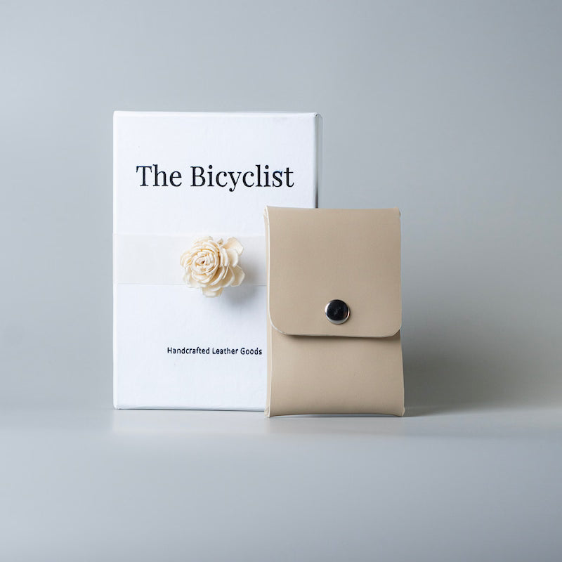 A off white leather card pouch with a snap button closure stands in front of a white gift box labeled 'The Bicyclist' on a light gray background.