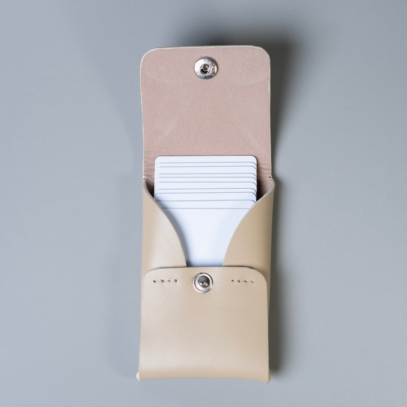 A off white full grain leather card holder wallet with silver snap buttons from The Bicyclist, shown open with white cards partially tucked inside, on a solid gray background.