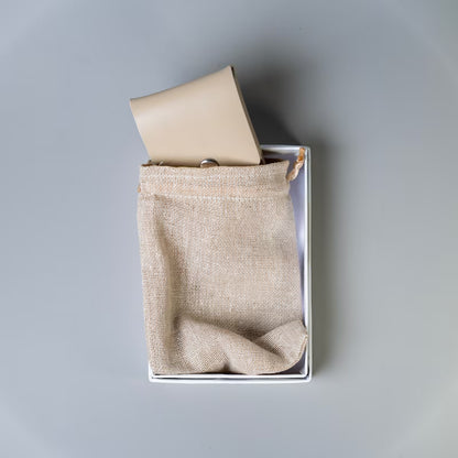 A off white full grain leather card holder or wallet with a snap button, partially tucked into a small beige linen drawstring pouch inside a paper cardboard gift box, on a neutral gray background.