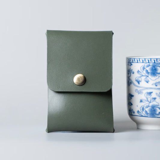 An olive green full grain leather business card holder or small card pouch with a snap button closure made by 'The Bicyclist', shown standing upright next to a blue and white ceramic cup on a light gray background.