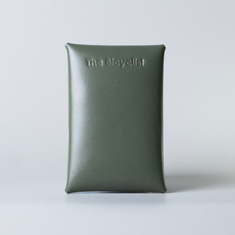 An olive green card pouch or business card case with the brand name 'The Bicyclist' embossed on the back, set against a plain light gray background.
