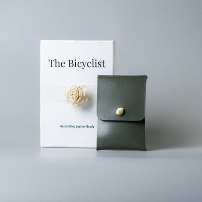 An olive green full grain leather card pouch or card holder with a gold snap button, displayed next to its white packaging box labeled 'The Bicyclist' on a neutral gray background.