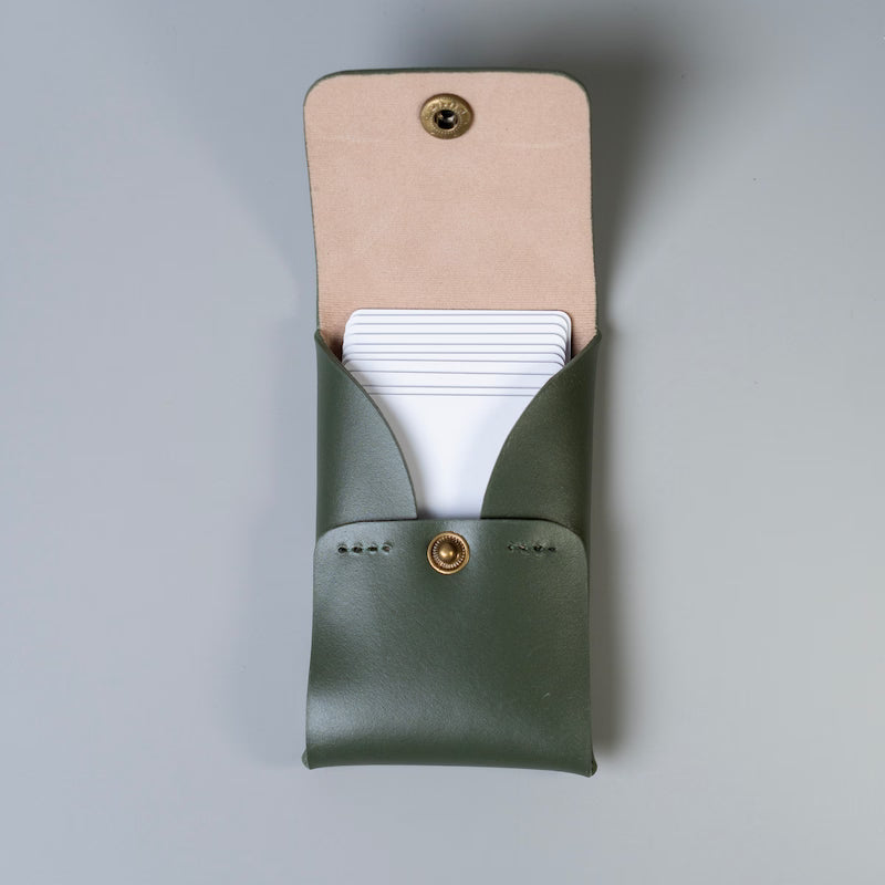An olive green full grain leather business card holder with a beige felt lined flap and a gold snap button shown open, made by 'The Bicyclist' with white plastic cards partially tucked inside, set against a neutral gray background.