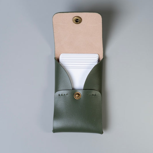 An olive green full grain leather business card holder with a beige felt lined flap and a gold snap button shown open, made by 'The Bicyclist' with white plastic cards partially tucked inside, set against a neutral gray background.