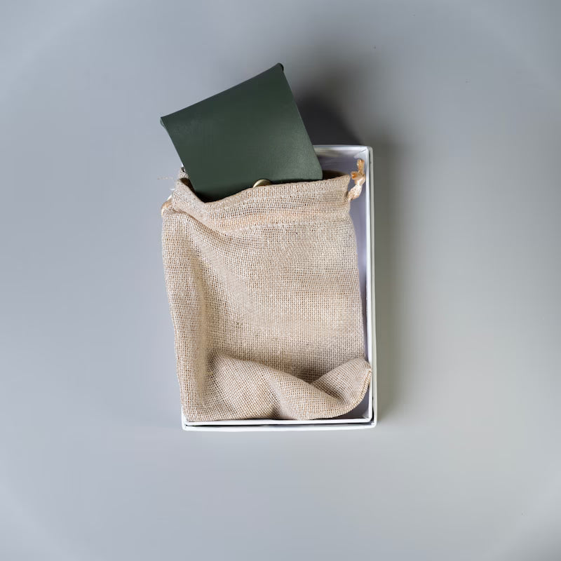 A olive green full grain leather card holder or card case made by 'The Bicyclist' half tucked inside a beige burlap drawstring pouch inside a white box, photographed from above on a light gray background.
