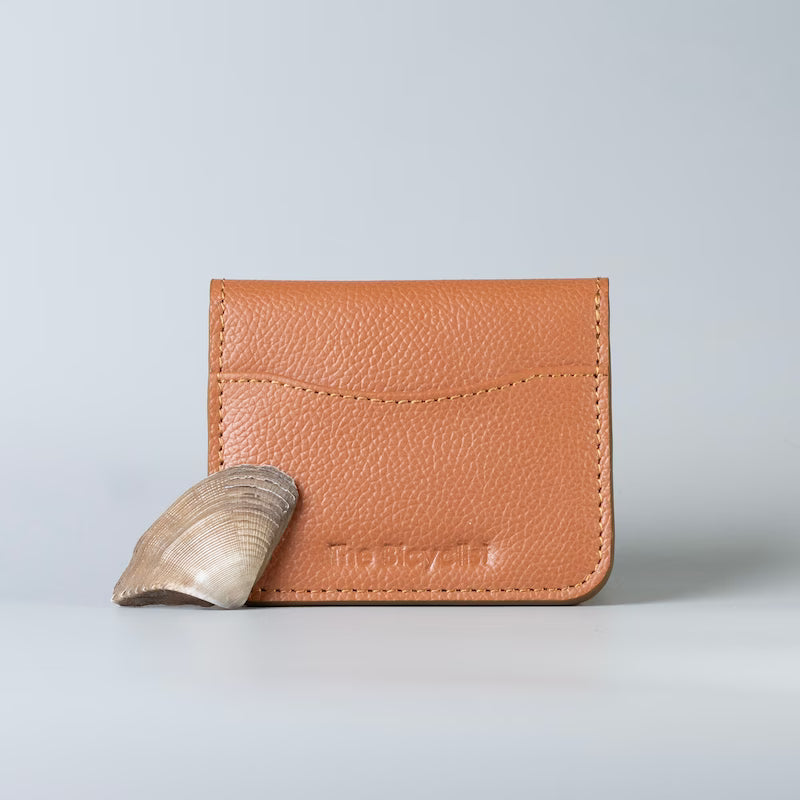 A tan pebbled leather card holder wallet with 'The Bicyclist' debossed on the front and a small sea shell leaning against it, set against a light gray studio background.
