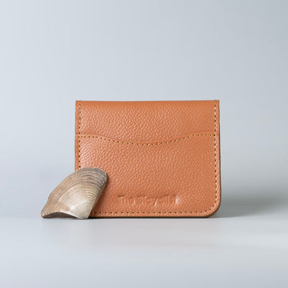 A tan pebbled leather card holder wallet with 'The Bicyclist' debossed on the front and a small sea shell leaning against it, set against a light gray studio background.