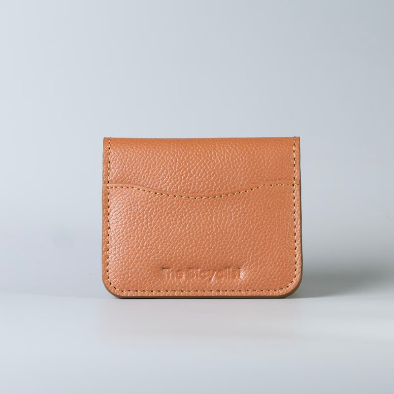 A small tan pebbled leather card holder wallet with a stitched front pocket and 'The Bicyclist' debossed logo, photographed on a light gray background.