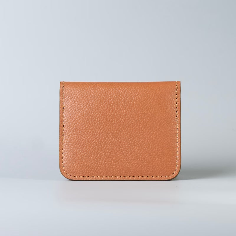 A small pebbled minimalist leather card holder or wallet in a warm tan/caramel color, photographed upright against a clean light gray studio background.