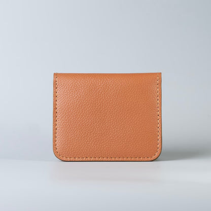 A small pebbled minimalist leather card holder or wallet in a warm tan/caramel color, photographed upright against a clean light gray studio background.