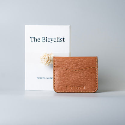 A tan pebbled leather card holder wallet with 'The Bicyclist' embossed on the front, displayed next to its white packaging box with a cream flower accent on a light gray background.