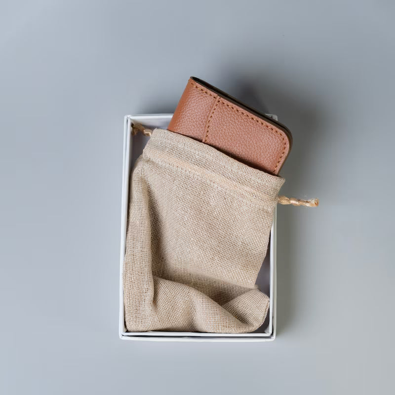 A brown pebbled leather wallet or card holder partially tucked into a white box with a beige drawstring dust bag, photographed from above on a light gray background.