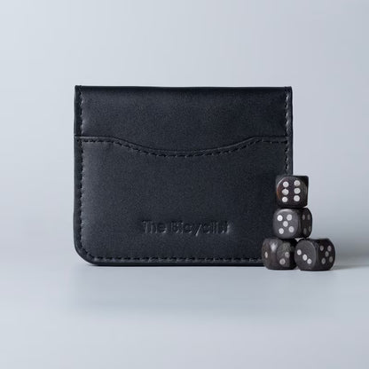 A black leather card holder wallet with visible stitching and debossed branding of The Bicyclist, shown on a light gray background with four dark dice stacked beside it.