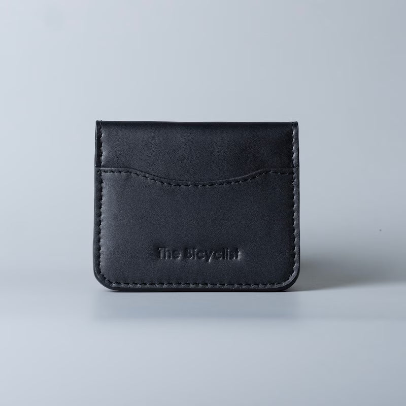 A black full grain leather card holder wallet with visible stitching and debossed 'The Bicyclist' logo, centered on a light gray background.