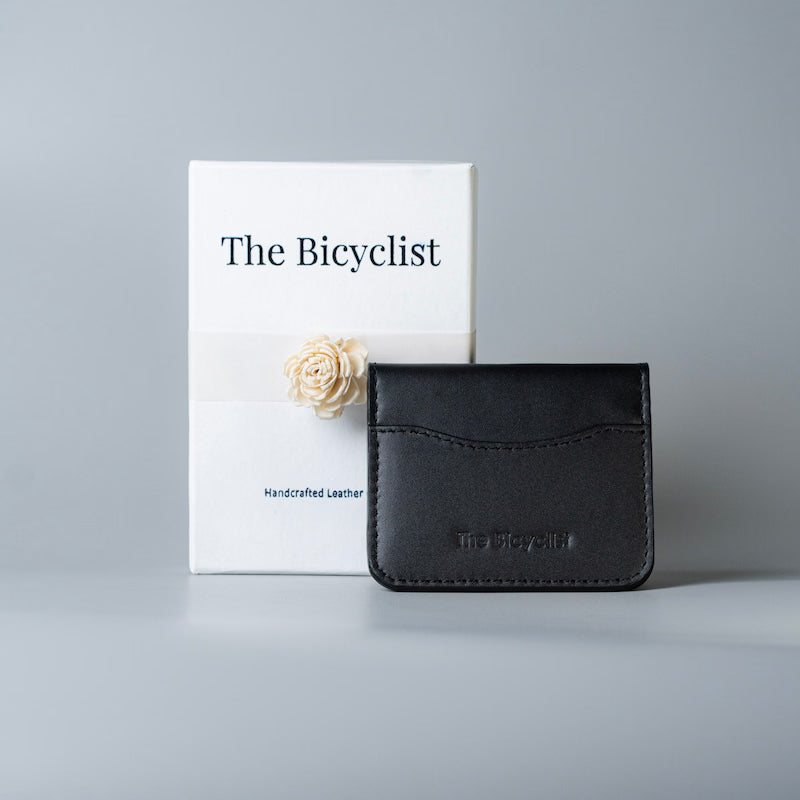 A black full grain leather card holder wallet with debossed branding, of 'The Bicyclist' displayed next to its white packaging box on a neutral gray background.