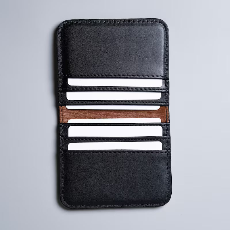 A black full grain leather card holder wallet with a tan sheep leather lining on the inside, shown open with six plastic cards in each of the card slot on a light gray background.