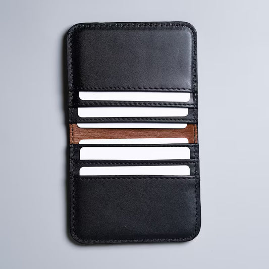 A black full grain leather card holder wallet with a tan sheep leather lining on the inside, shown open with six plastic cards in each of the card slot on a light gray background.