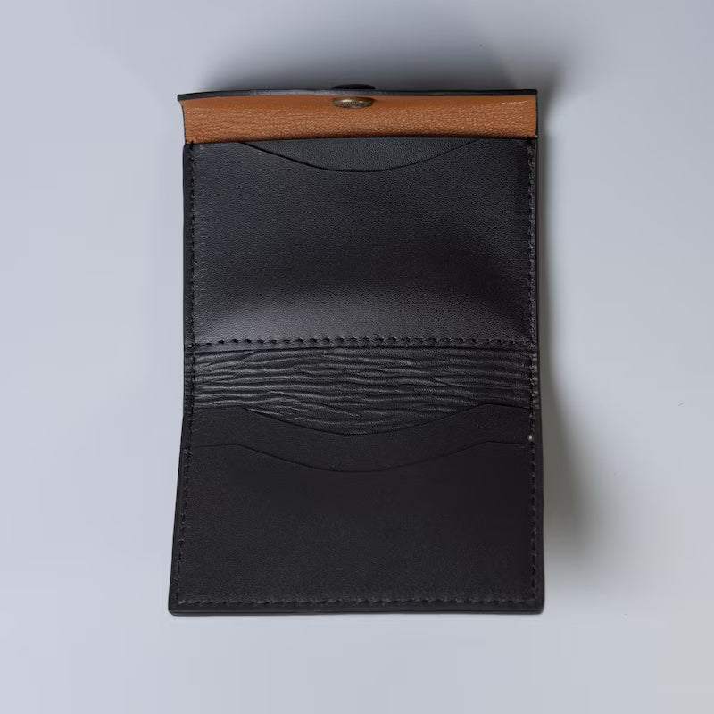 A minimalist black full grain leather card holder wallet with a tan sheep leather lining on the inside from 'The Bicyclist', shown open against a light gray background.