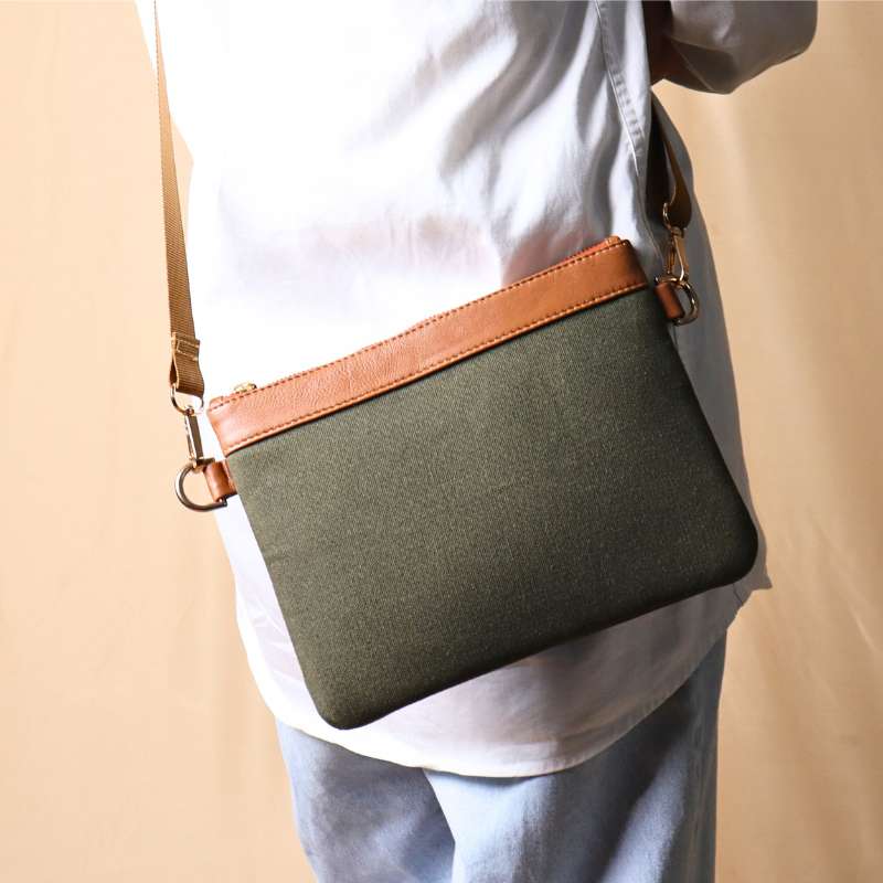 Back View of Woman in a white shirt and light blue denims carrying over her shoulder, a dark green canvas crossbody sling bag with tan leather trims on the top and green nylon strap.