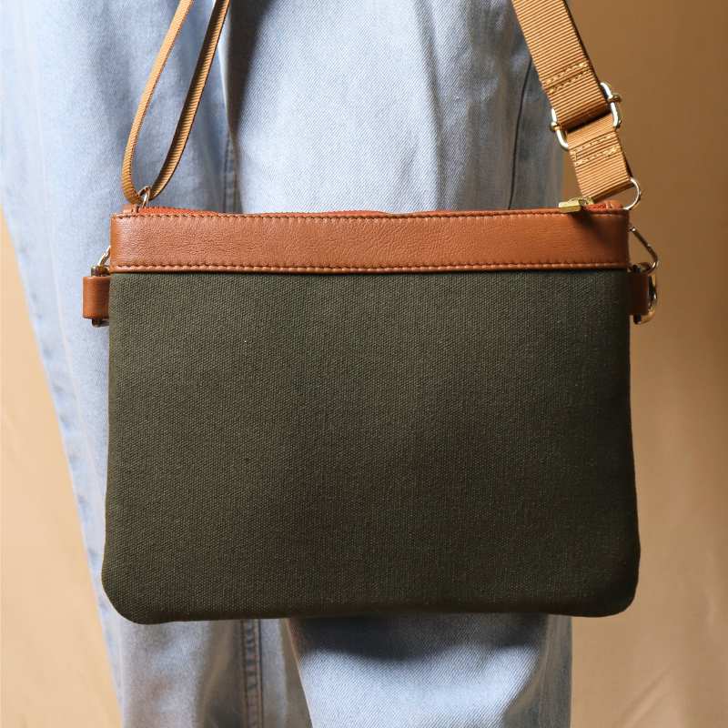 Woman in light blue denims carrying a dark green canvas crossbody sling bag with tan leather trims on the top and green nylon strap.