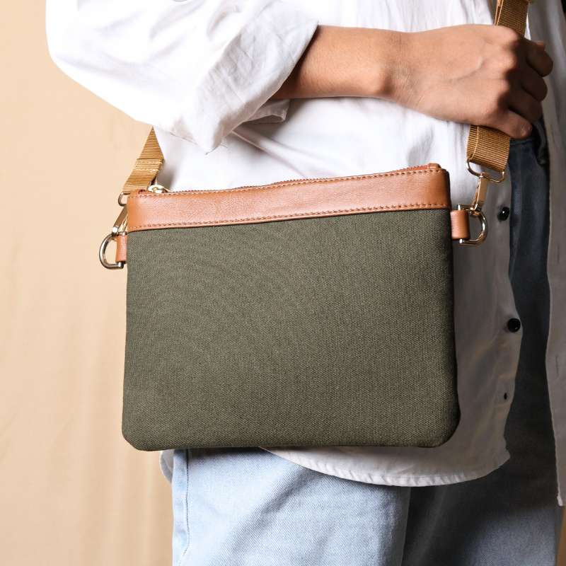 Woman in a white shirt and light blue denims carrying over her shoulder, a dark green canvas crossbody sling bag with tan leather trims on the top and green nylon strap.