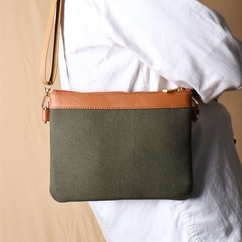 Side View of Woman in a white shirt and light blue denims carrying over her shoulder, a dark green canvas crossbody sling bag with tan leather trims on the top and green nylon strap.