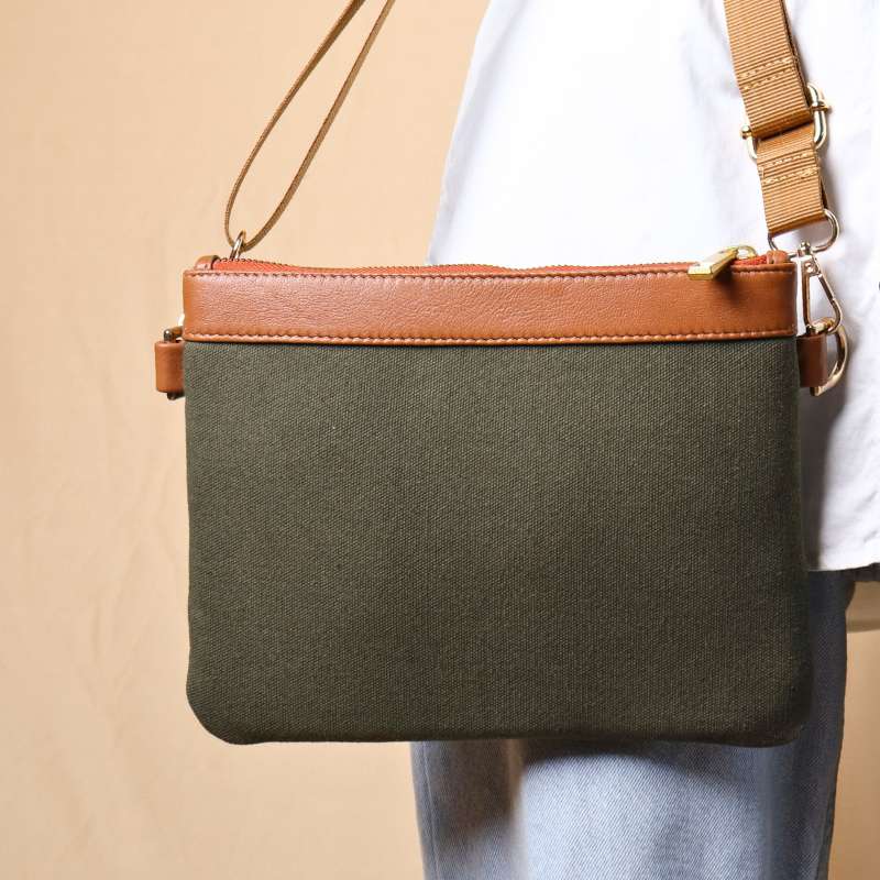 Woman in light blue denims carrying a dark green canvas crossbody sling bag with tan leather trims on the top and green nylon strap.