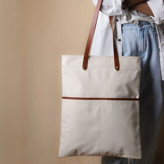 Woman in a white shirt and light blue denims holding in her hand, a off-white canvas tote with leather straps and leather trim front pocket
