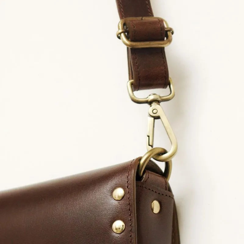 Detail Strap Fittings View of a Dark Brown Leather Crossbody Sling Bag with a Flap and Light Gold fittings on a white background