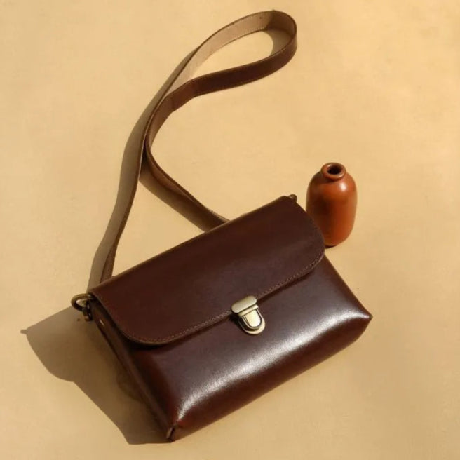 Dark Brown Leather Crossbody Sling Bag with a Flap and Light Gold Pushlock and fittings with a small terracotta vase & bottles of perfume on a light beige background
